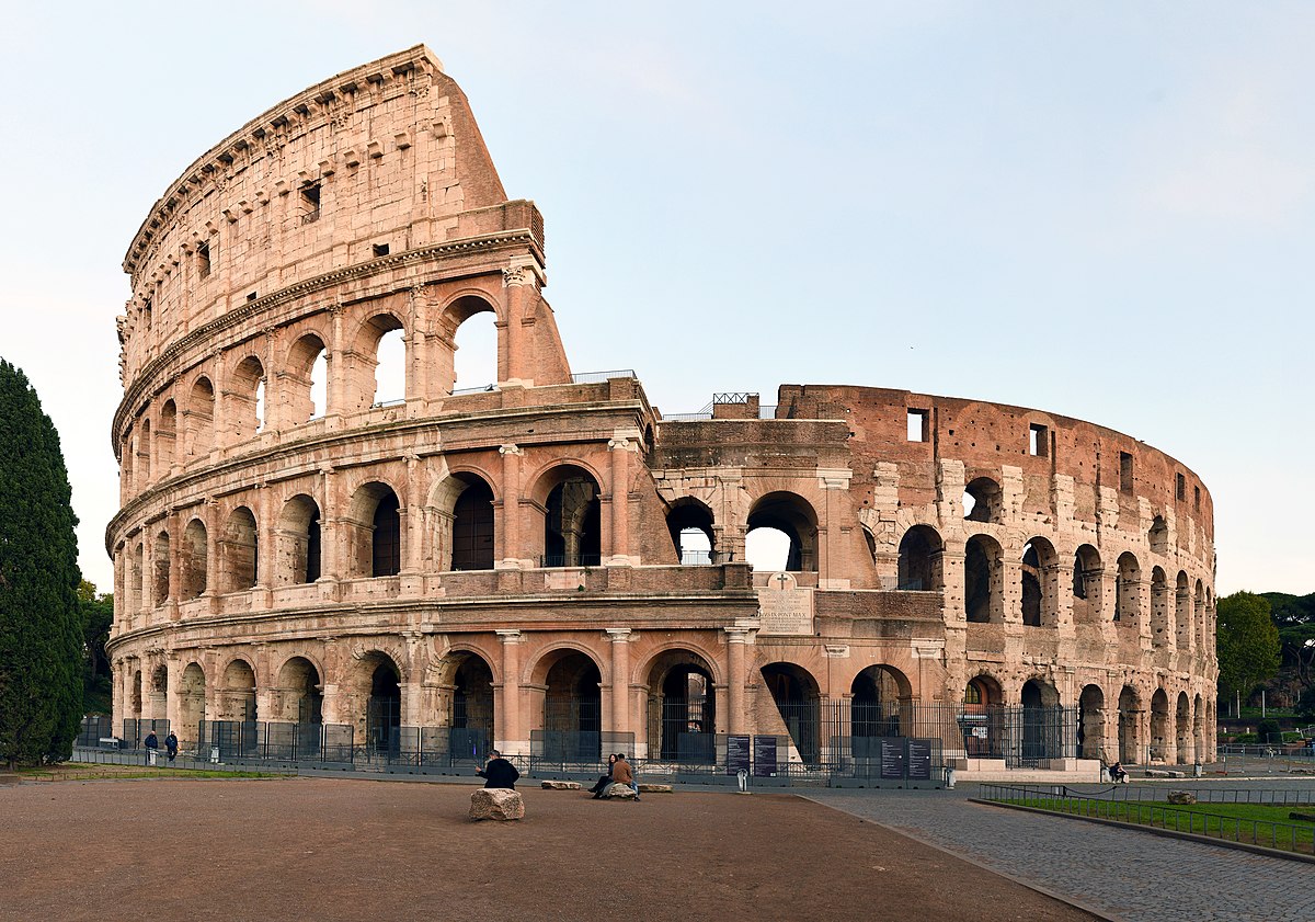 The circular 4-storey round building made of stone looks like it has fallen in half and at the same time people are watching it sitting on the stone.
