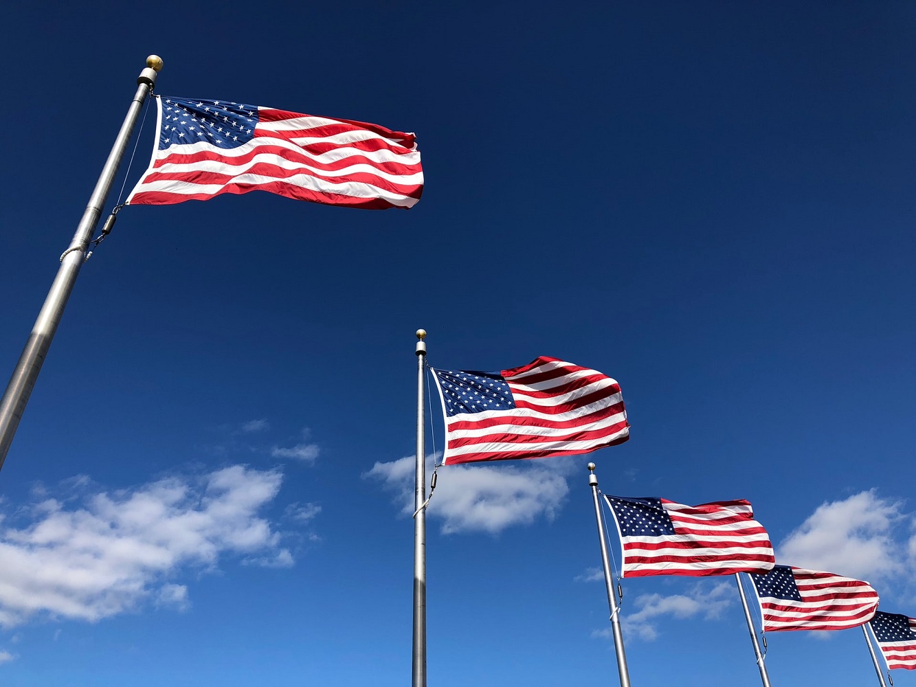 Five American flags in metal poles waving amidst a blue sky with some clouds
