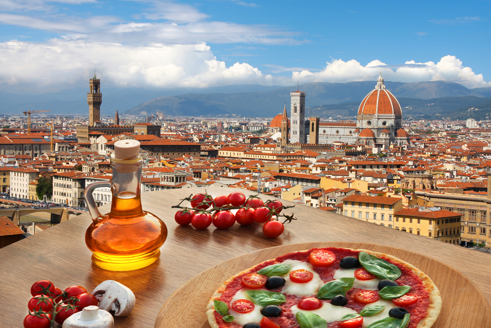 A pizza and some ingredients on the table with the city view in the backdrop