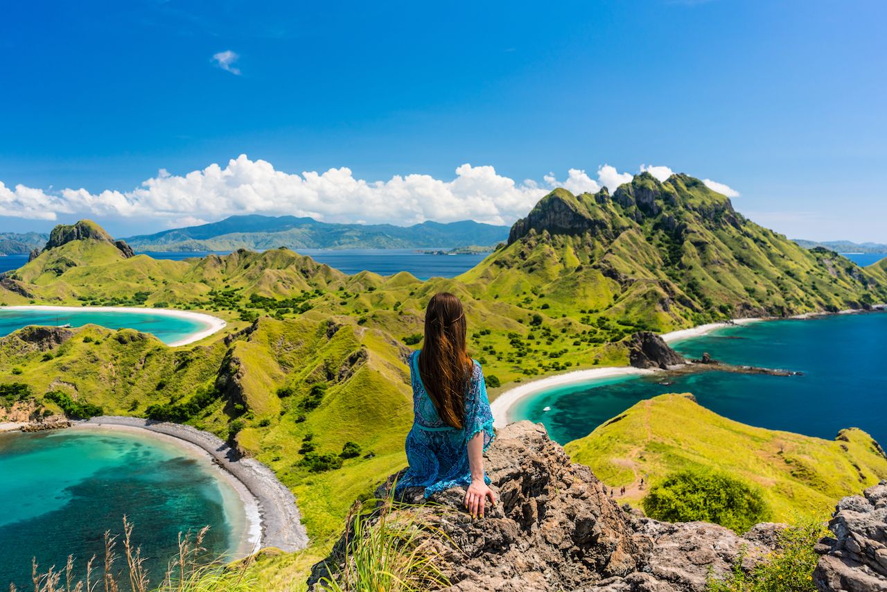 In the middle, a woman is sitting in a stone and looking out at the beautiful bodies of water and the mountains in front of her