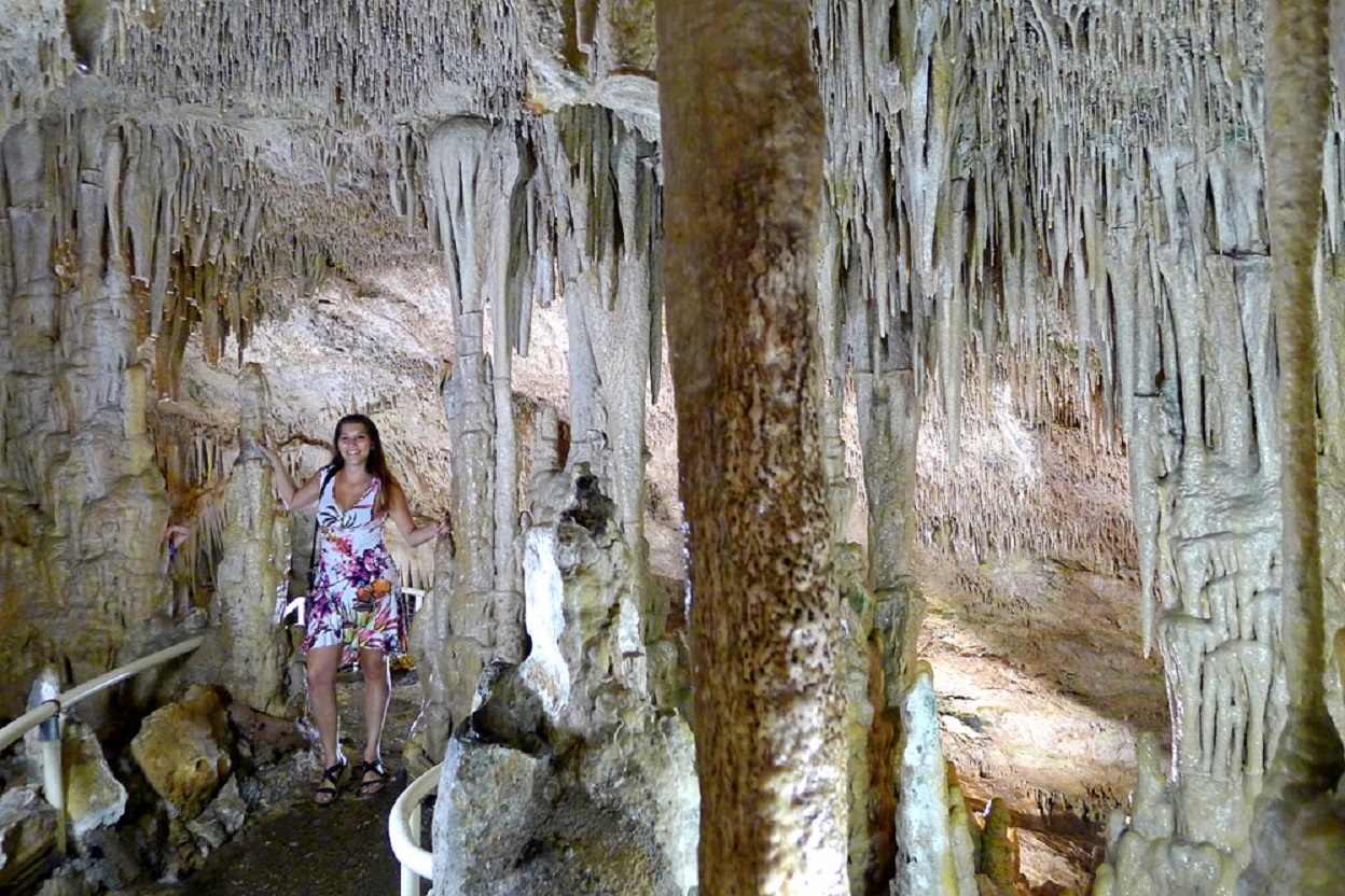 A smiling adult woman in a white dress with floral prints inside a cave with stalactites in Cote d’Azur